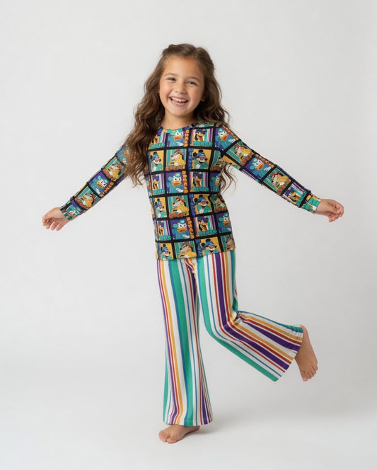 A young girl with long brown hair is smiling and posing in a colorful two-piece pajama set. The set features a long-sleeve top with a cartoon character grid pattern and matching wide-leg pants with vertical rainbow stripes, set against a plain light gray studio background.