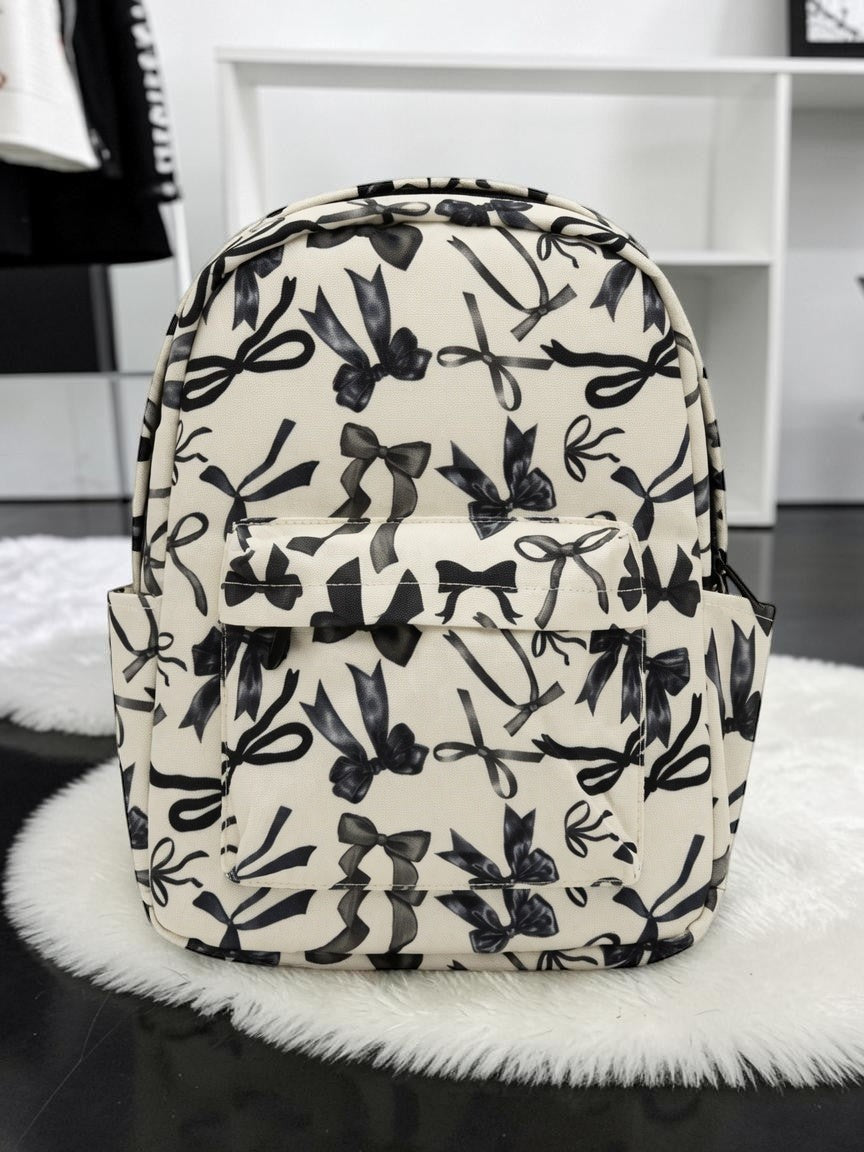 Little Britches Children's Boutique cream mini backpack with a black bow print, featuring a front pocket and side slots.